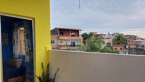A balcony with a view of a residential area, a plant, and a partially visible room with a bed and curtains. - House, 2 bedrooms with air conditioning, airy, fully equipped. (Salvador)