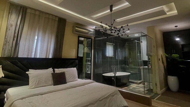 A modern bedroom with a large bed, a glass shower enclosure, and a contemporary ceiling design. - Pine Crest Hotel and Suites  (Abuja)