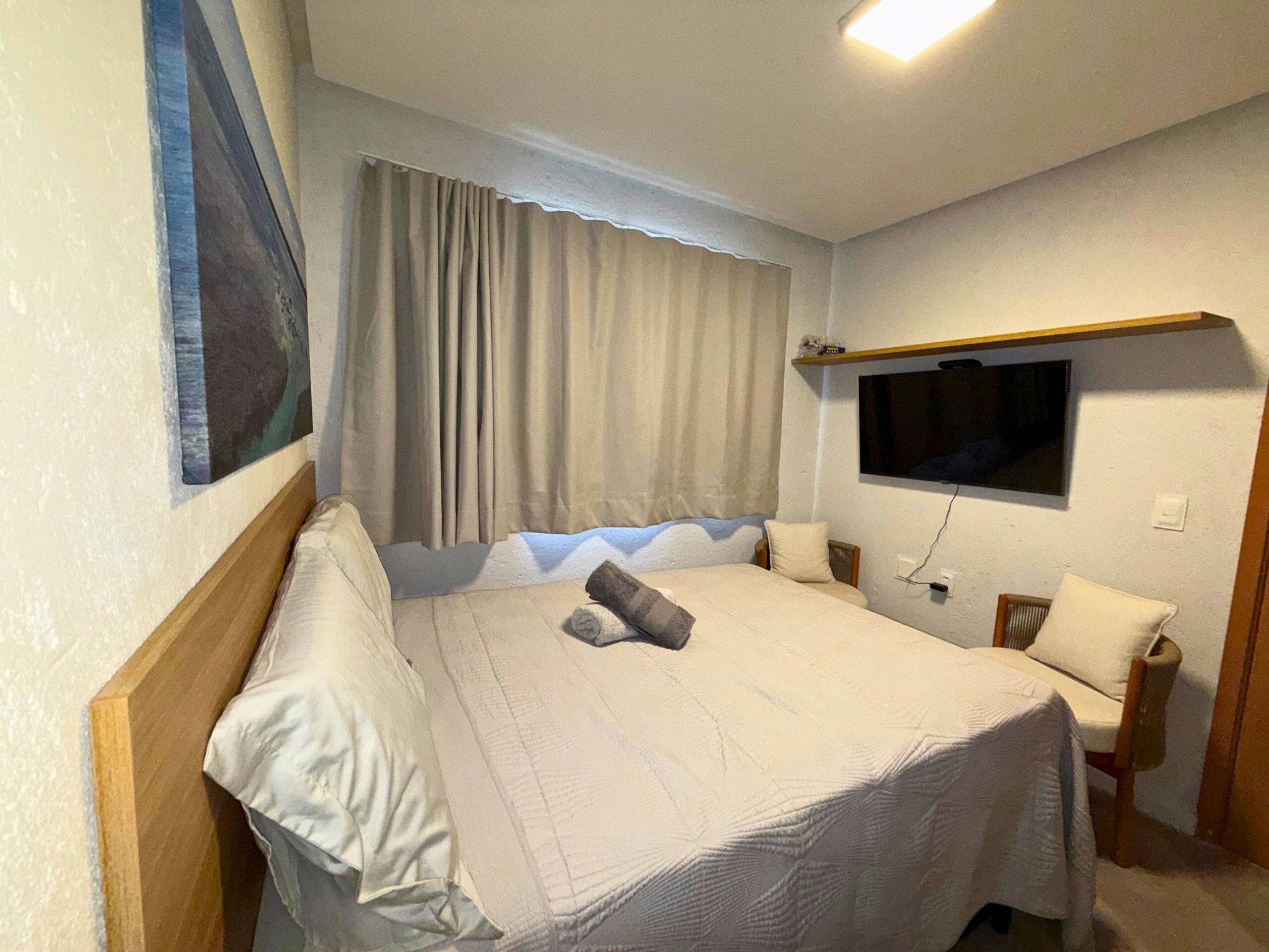 Habitación de hotel con cama, televisión montada en la pared, ventana con cortinas y un cuadro en la pared.