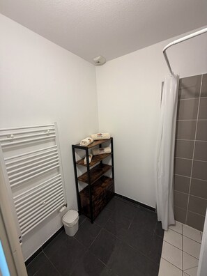 A small bathroom with a white radiator, a wooden shelf with towels, a toilet, and a shower area. - Biscarrosse apartment in town center, 10 minutes from the ocean and 5 minutes from the lake (Biscarrosse)