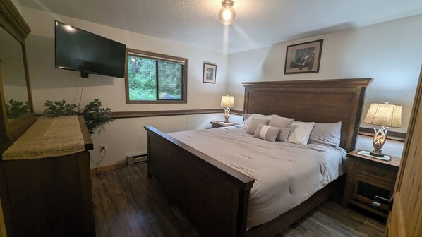A bedroom with a large bed, a TV mounted on the wall, a window with a view of greenery, and two bedside lamps. - New Listing! 3BR/2BA Ptarmigan Village w/Pool - Hotub - Sauna - Lake & Ski! (Whitefish)
