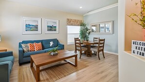 Un salón con un sofá azul, una mesa de centro de madera y una zona de comedor con una mesa y sillas de madera. Hay cuadros en la pared, una planta y un cartel de bienvenida.