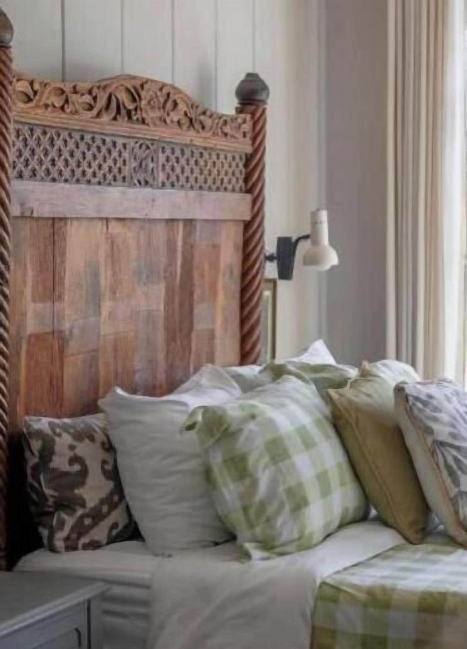 A bedroom with a wooden headboard, a bed with multiple pillows, a bedside table, and a window with curtains.
