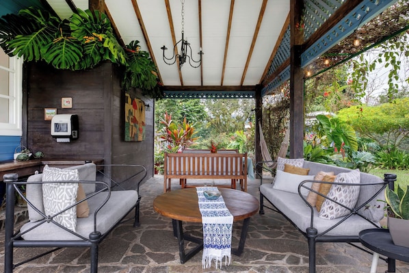 An outdoor seating area with a wooden bench and two sofas, a patterned table runner, and a hanging light fixture.
