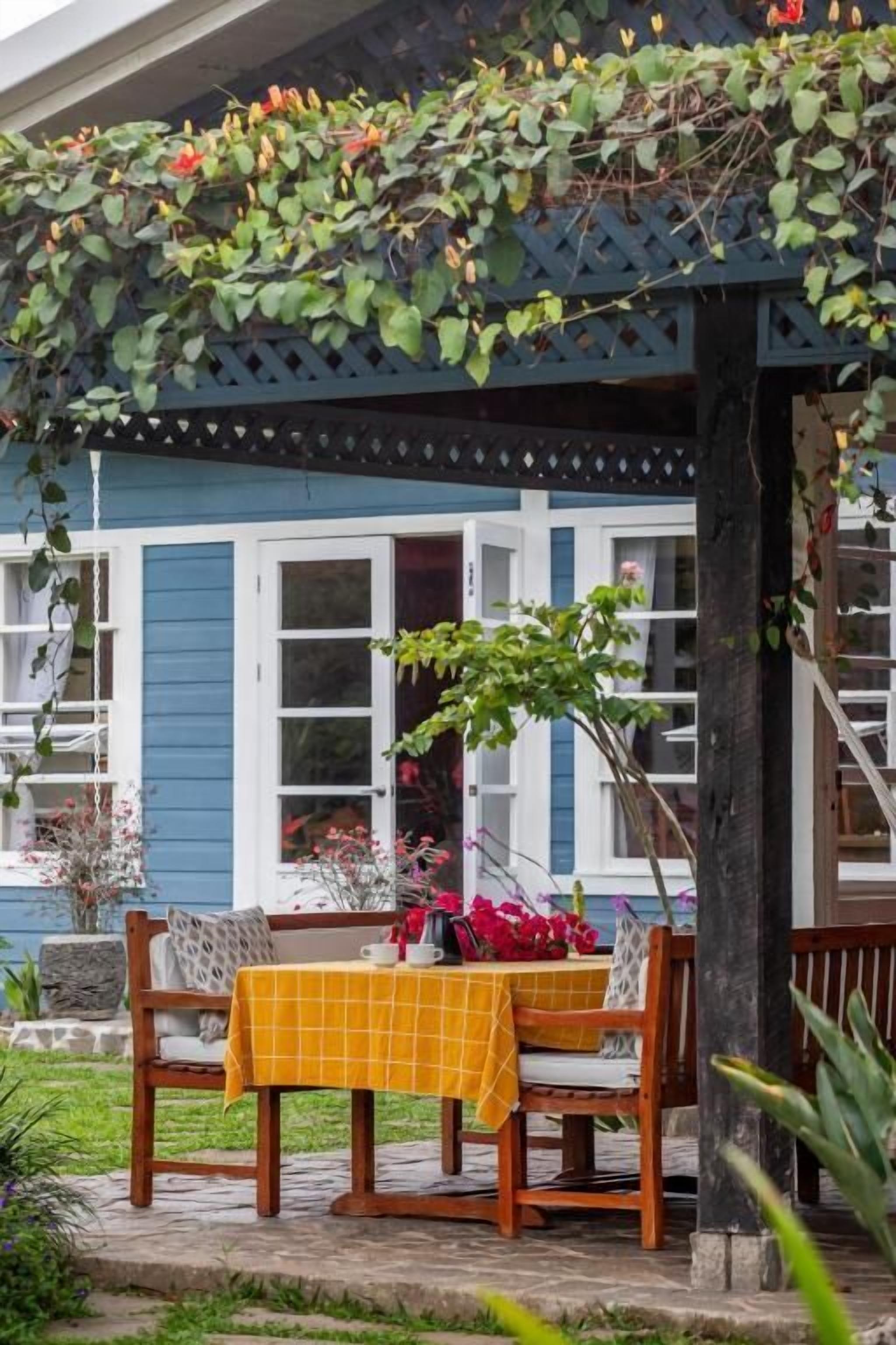 A covered outdoor seating area with a table and chairs, a wooden bench, and a lattice structure with climbing plants.