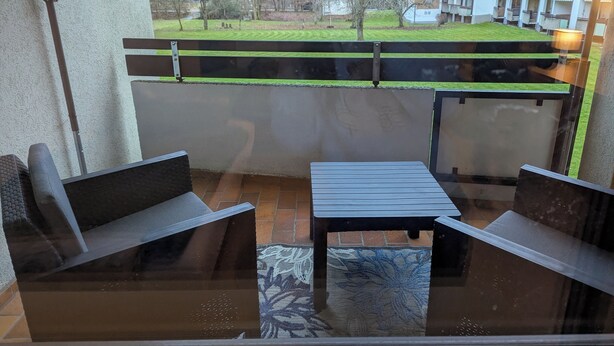 Ein Balkon mit Tisch und zwei Stühlen, Blick auf eine Wiese und Gebäude im Hintergrund.