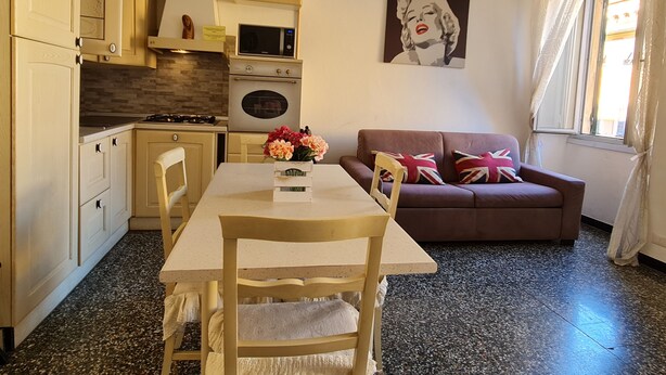 En kompakt opholdsstue med tekøkken, spisebord med stole, sofa med puder og et billede på væggen.