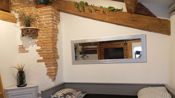 Une pièce chaleureuse avec un mur en briques, une poutre en bois au plafond, un lit avec un matelas noir et des coussins à motifs, une petite armoire blanche et un miroir au mur.