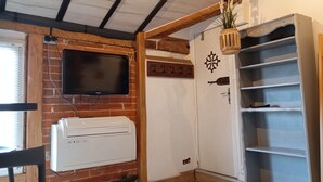 Une pièce avec un mur en briques, un téléviseur fixé au mur, un climatiseur blanc, une porte en bois ornée d’une plaque décorative et une étagère blanche.