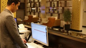 A man working at a reception desk with a computer, surrounded by a modern interior with a patterned wall and a decorative plant. - Aris in Apartment (Riyadh)