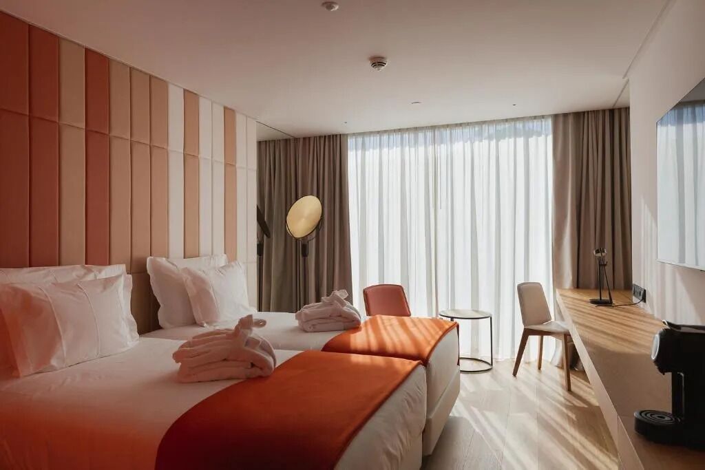 Een hotelkamer met twee bedden, een zithoek met een stoel, een klein tafeltje en een groot raam met gordijnen.