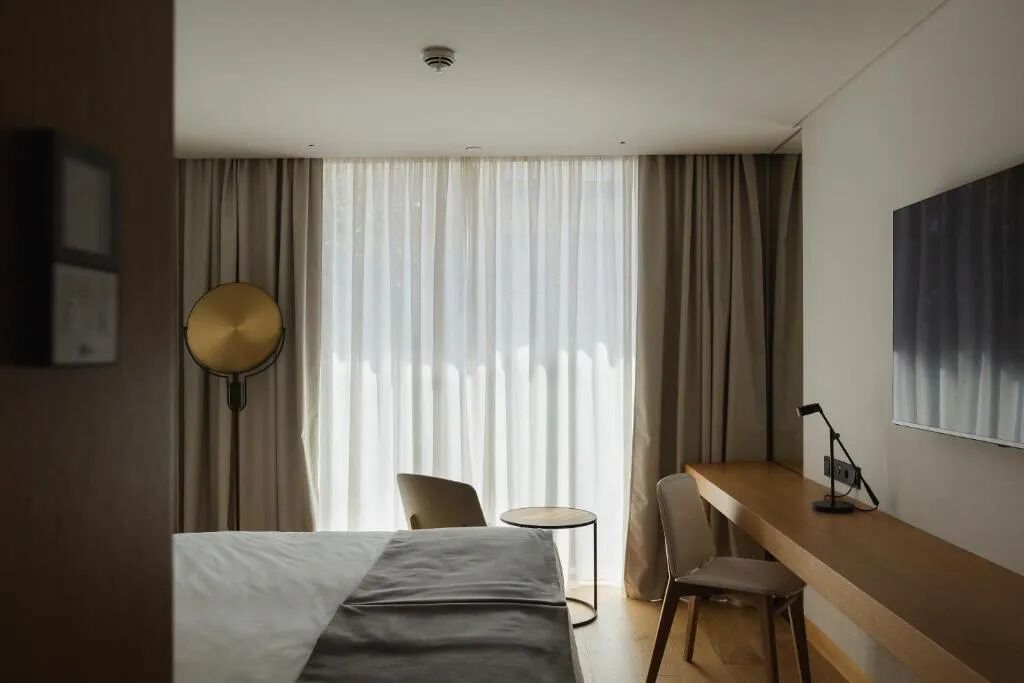 A hotel room with a bed, a desk, a chair, a lamp, and a large window with curtains.