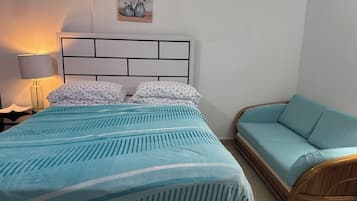 Een bed met een blauw-wit gestreept dekbed, een rieten bank, een nachtkastje met een lamp, en een aan de muur hangend schilderij.