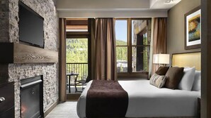 Habitación de hotel con una cama grande, un televisor montado en una pared de piedra, una chimenea y un balcón con mesa y silla.