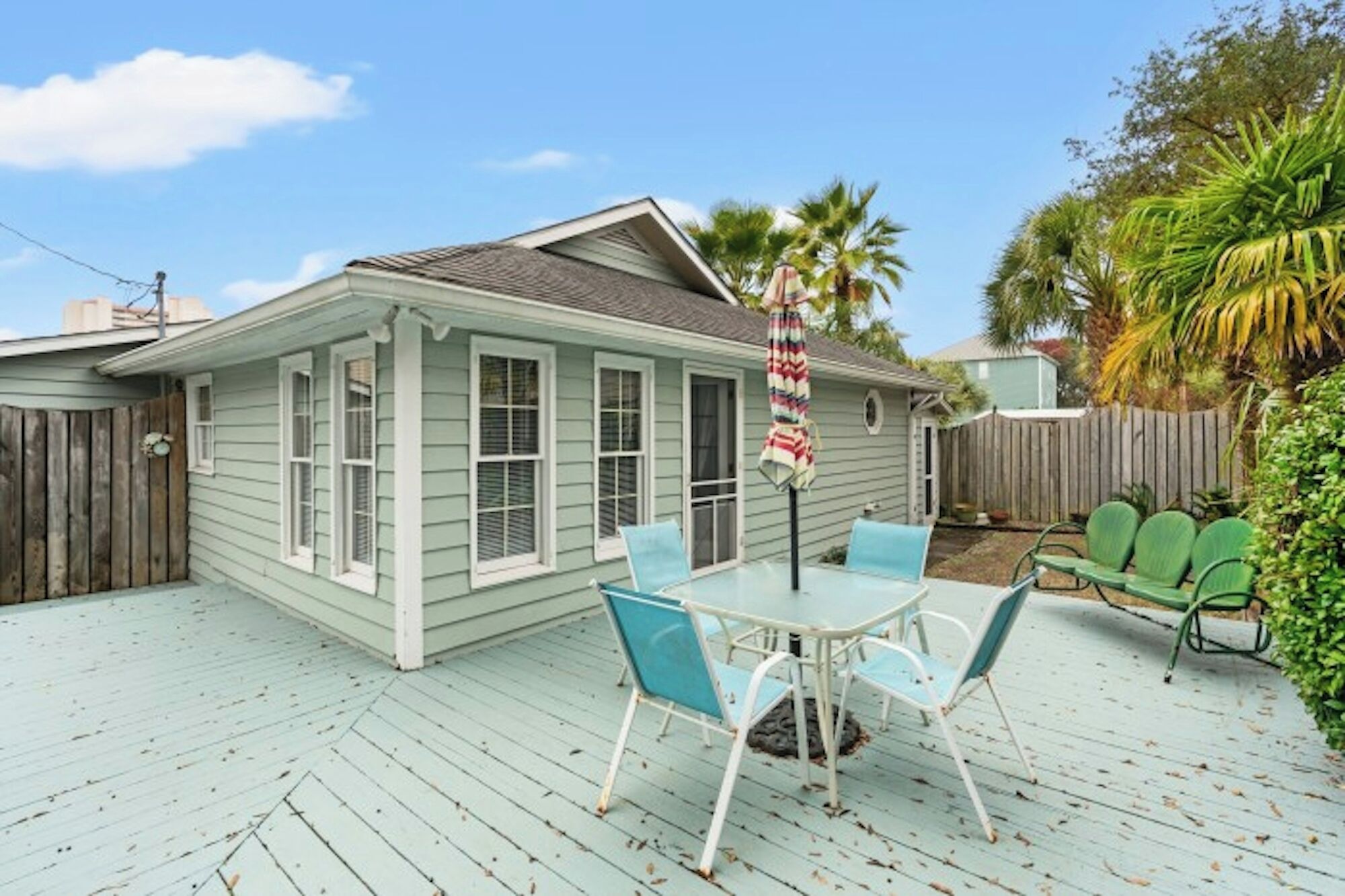 A house with a covered patio area featuring a table and chairs, a wooden deck, and a fenced backyard with palm trees.