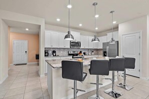 A modern kitchen with white cabinets, a central island with bar stools, stainless steel appliances, and pendant lighting. - 5BD Windsor Island Resort Disney Themed (WI 3858) (Davenport)