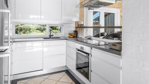 Une cuisine moderne avec des armoires blanches, une hotte de cuisinière en acier inoxydable, un four encastré et une fenêtre donnant sur la verdure à l’extérieur.
