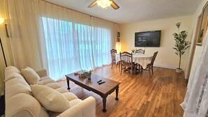 A living room with a sofa, a coffee table, a dining table with chairs, a ceiling fan, and a TV mounted on the wall. - Ami's Oak House - ORNL - Melton Lake - Windrock (Oak Ridge)