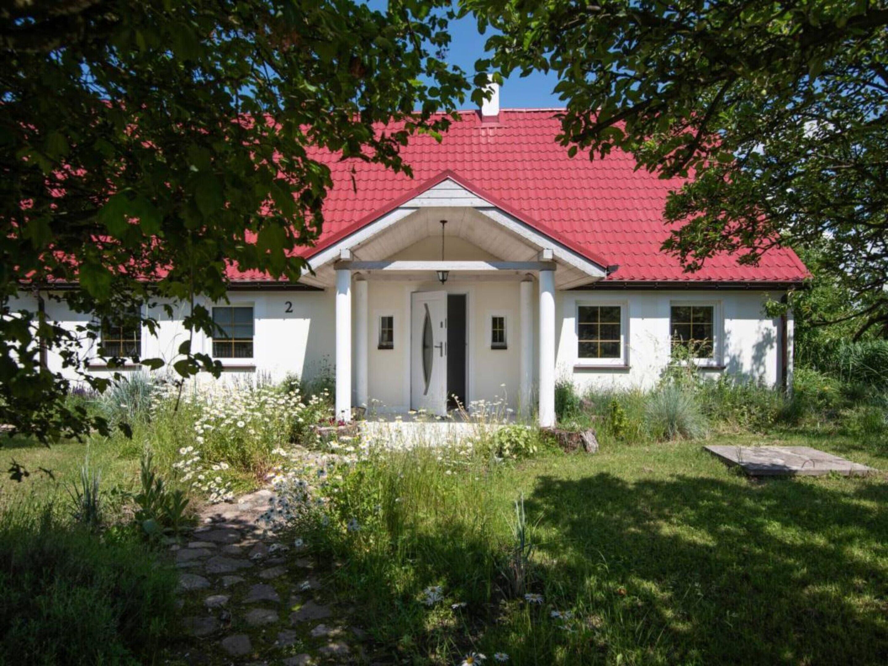 A white building with a red roof, a stone pathway leading to the entrance, and a garden with various plants and flowers.