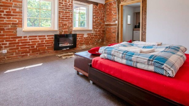 A bedroom with exposed brick walls, a fireplace, a bed with red bedding, and two windows. - Comfortable House With a Swimming Pool and Sauna (gmina Dobra)