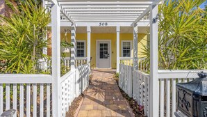 A covered walkway leading to a yellow building with the number 508, flanked by white picket fences and palm trees.