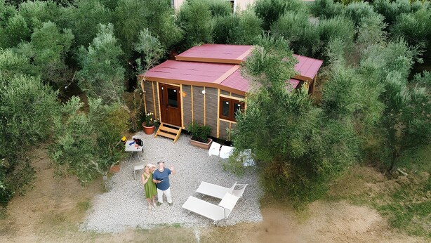Una cabaña rodeada de vegetación densa, con un sendero de grava que lleva a una terraza de madera y un pequeño espacio al aire libre con mesas y sillas.