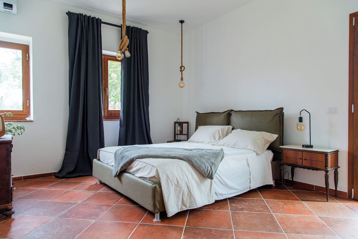A bedroom with a bed, a bedside table, a lamp, a hanging light, and a window with curtains.