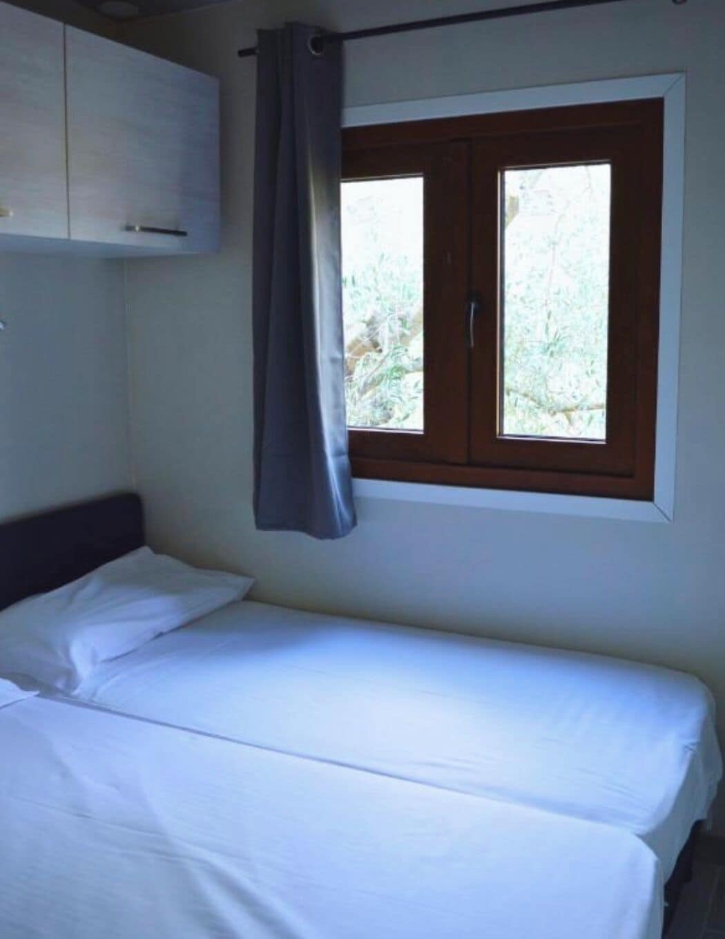 Ein Einzelbett mit weißer Bettwäsche, ein Holz-Kopfstück, ein Fenster mit Blick auf Grünflächen und ein weißer Kleiderschrank.