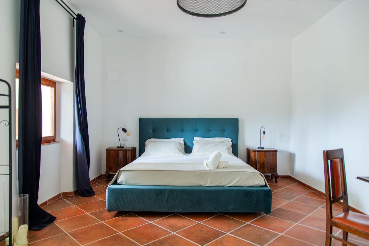 A bedroom with a large bed, two bedside tables, a chair, a window with curtains, and a ceiling light fixture.