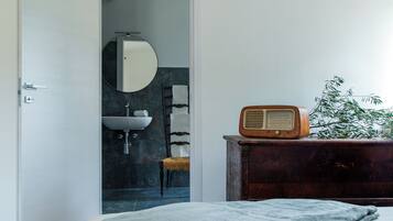 Un dormitorio con cama, cómoda, radio, planta y baño visible a través de una puerta abierta.