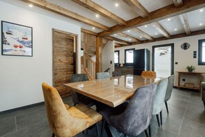 A dining area with a wooden table and upholstered chairs, a staircase, and a kitchen area. - Le Gypaète, Prodains, Morzine (Morzine)