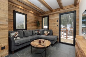 A modern interior with a sectional sofa, a round wooden coffee table, and a view of a snowy outdoor deck. - Le Gypaète, Prodains, Morzine (Morzine)