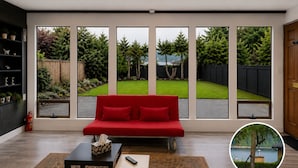 Une pièce avec un canapé rouge, une petite table sur laquelle se trouvent une boîte de mouchoirs et une télécommande, et un tapis. De grandes fenêtres offrent une vue sur un jardin et un lac.