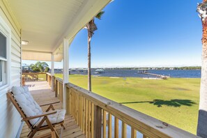 A wooden deck with a white cushioned chair, a view of a lake, and a dock extending into the water. - La Flor: Beautiful and serene water front oasis. Easy beach access! (Gulf Shores)