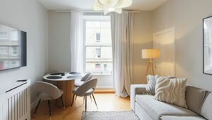 A modern living room with a sofa, a dining table with chairs, a lamp, and a large window with curtains. - Cosy & Stylish Edinburgh City Flat - Pass the Keys (Edinburgh)