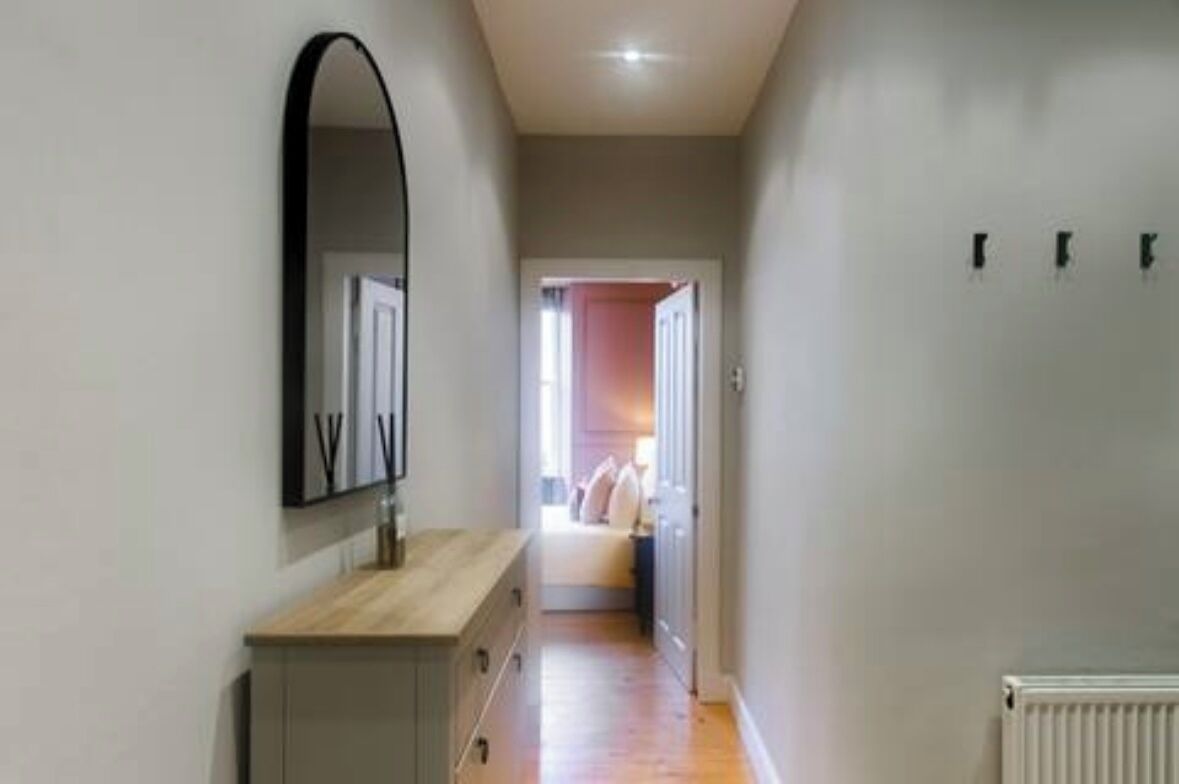 A narrow hallway with a wooden dresser and a mirror on the left, leading to a bedroom with a bed and a nightstand.