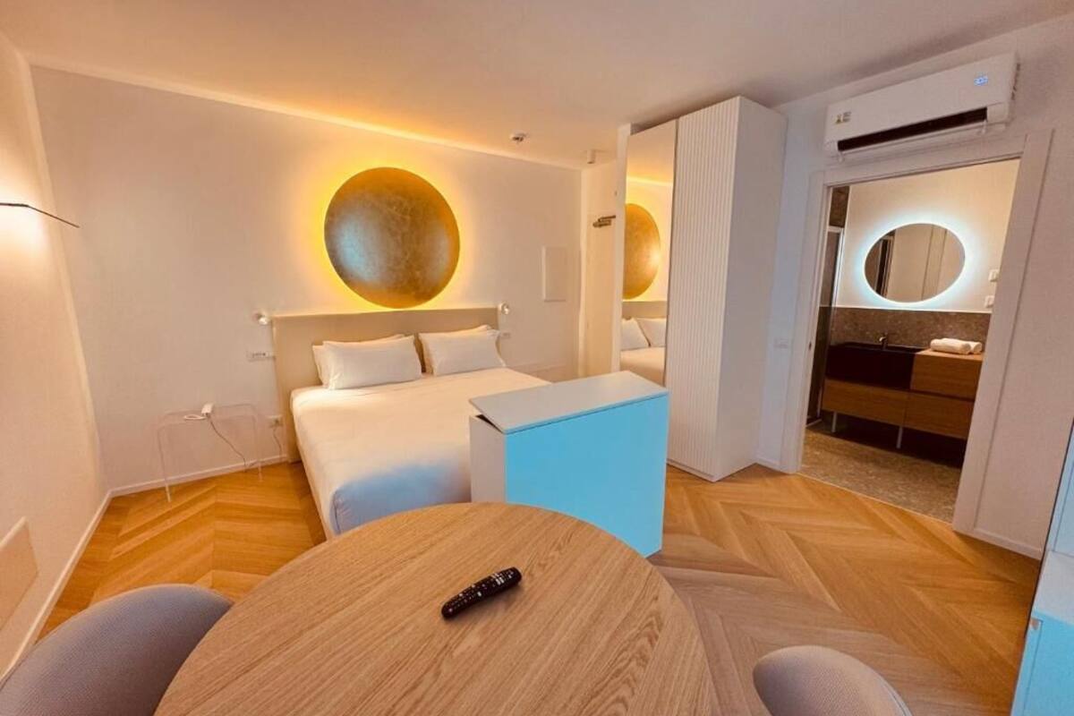 A modern hotel room with a large bed, a round wall light, a small table, and a bathroom with a mirror and sink.