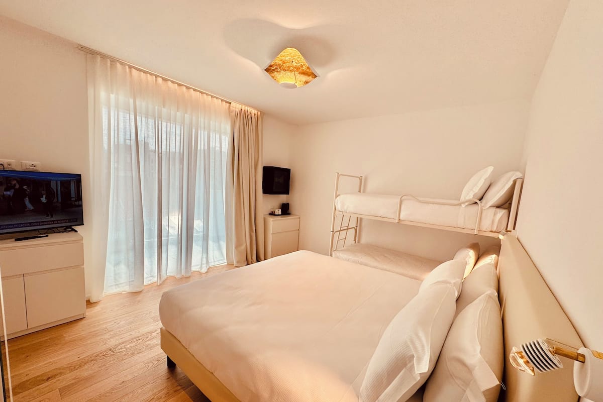 A hotel room with a large bed, a TV on a bedside table, a desk with a computer, and a window with sheer curtains.