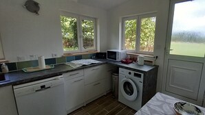 Een compacte keuken met een wasmachine, een magnetron en een spoelbak. Twee ramen zorgen voor natuurlijk licht en uitzicht op groen.
