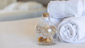 A clear glass bottle with dried flowers, a rolled white towel, and a bed in the background.