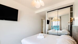 A modern hotel room with a large bed, a television mounted on the wall, and a large mirror reflecting the room and a balcony with a view of the sea.