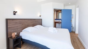 A modern bedroom with a wooden headboard, a bedside table with a remote control, and a neatly made bed with rolled towels. - Au pied de la plage 1 - Vue mer (Cancale)