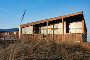 A wooden building with large windows and a balcony, surrounded by tall grass. - Brouwersdam Stay (Ouddorp)