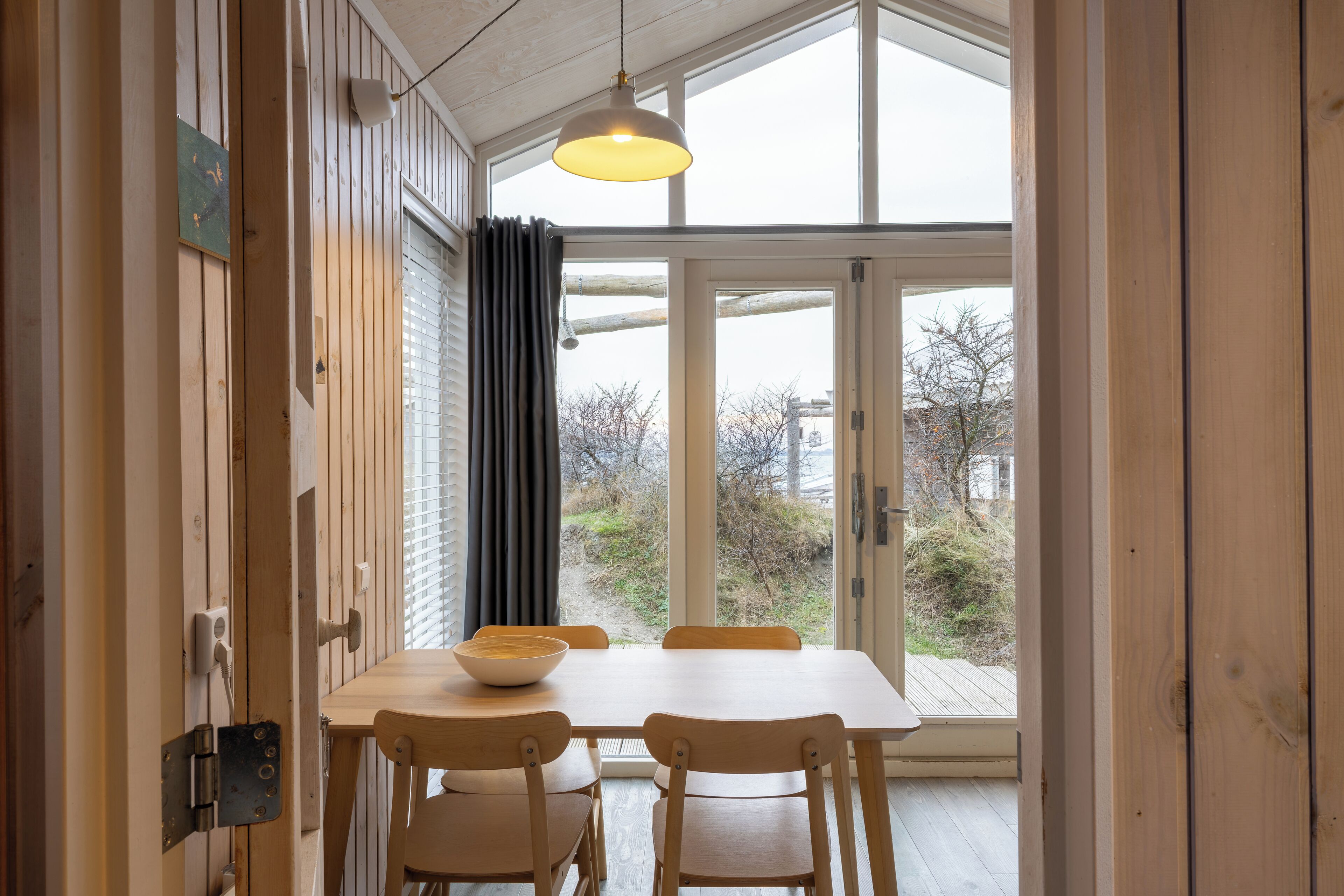 A wooden dining table with chairs, a bowl, and a pendant light in a room with large windows and a view of trees and buildings outside.