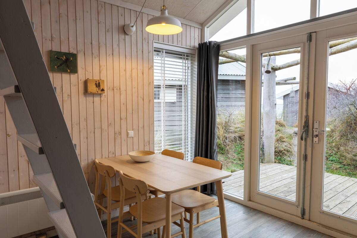 A dining area with a wooden table and chairs, a hanging light fixture, and a view through a glass door to an outdoor deck.