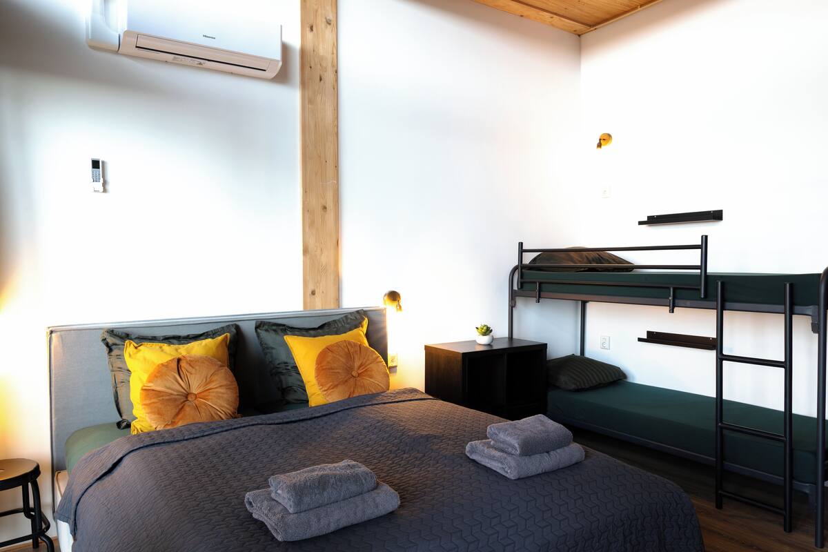 A room with a bunk bed, a bedside table with a lamp, and a bed with a patterned blanket and pillows.