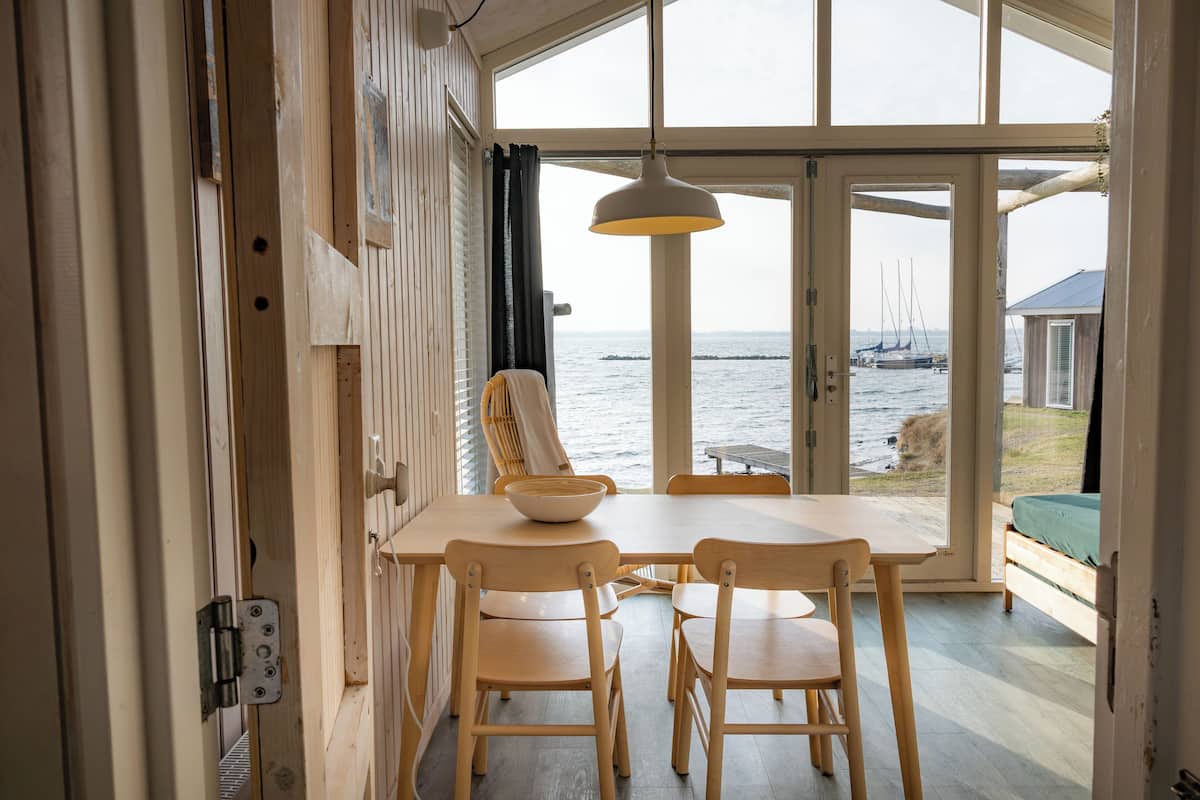 A wooden dining table with chairs, a hanging light fixture, and a view of the sea through large glass doors.