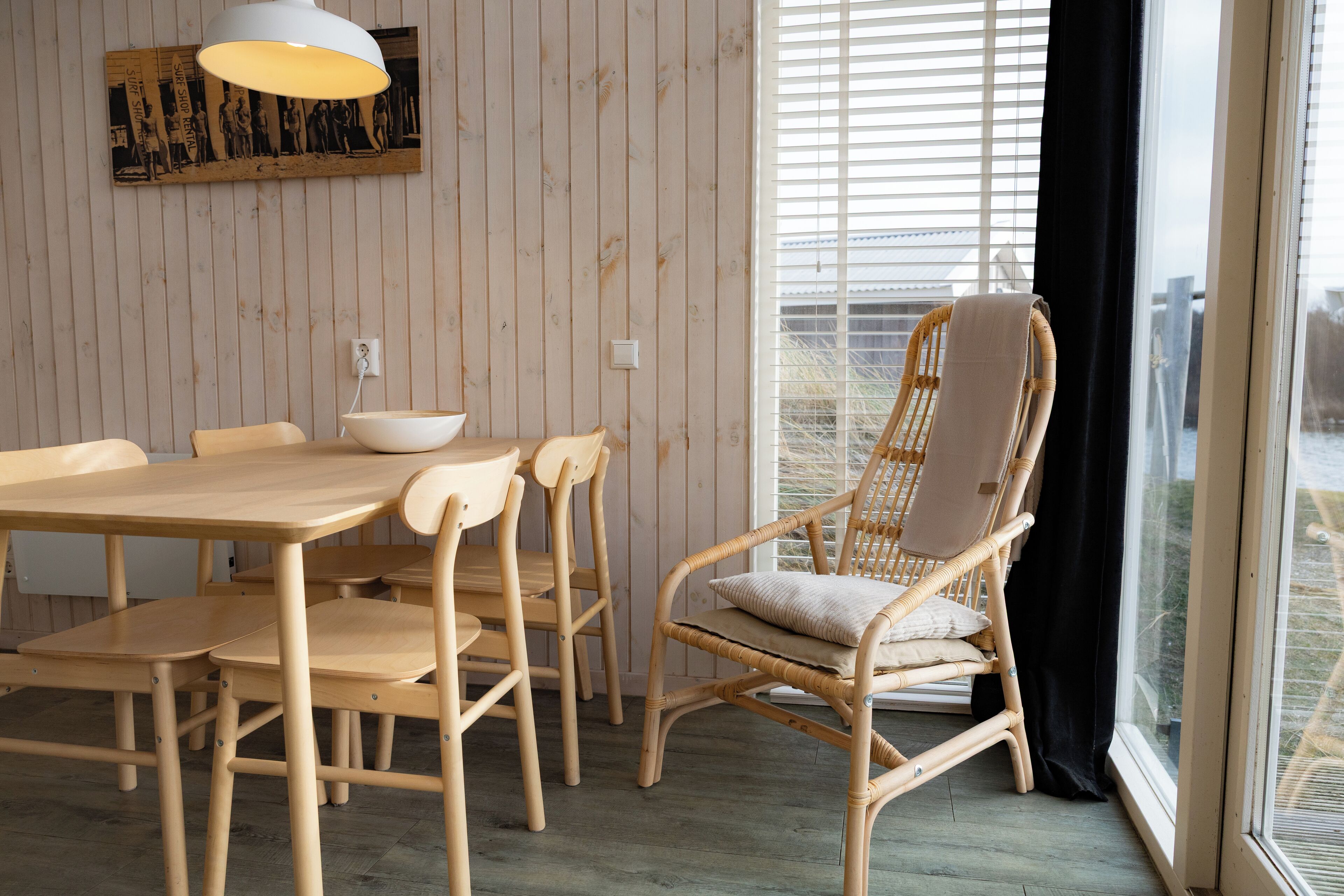 A dining area with a wooden table and chairs, a wicker chair with cushions, and a large window with blinds.