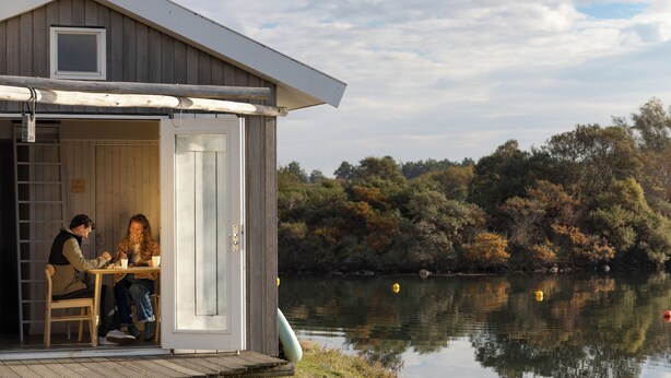 A wooden cabin with a door open, a table inside, and a view of a lake with trees. - Brouwersdam Stay (Ouddorp)