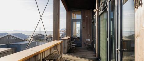 Una terraza de madera con mobiliario exterior, ventanas amplias y vistas a otros edificios.